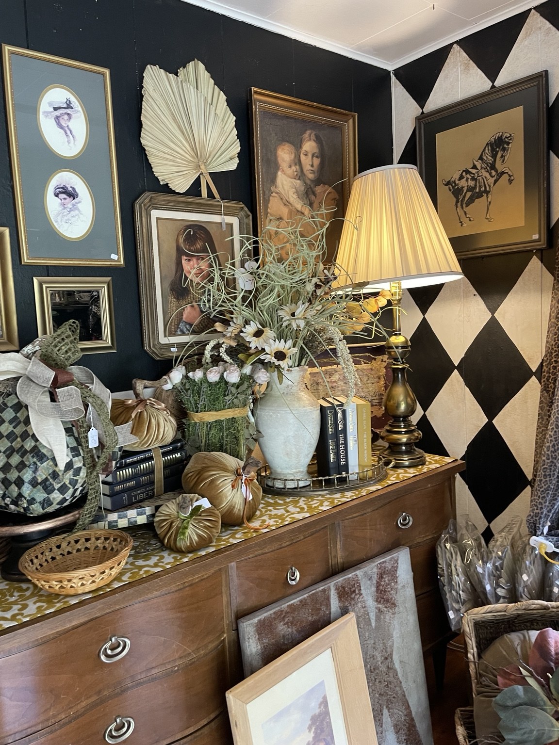 A wooden dresser styled with framed portraits, a glowing brass lamp, flowers, and pumpkins beside a harlequin wall